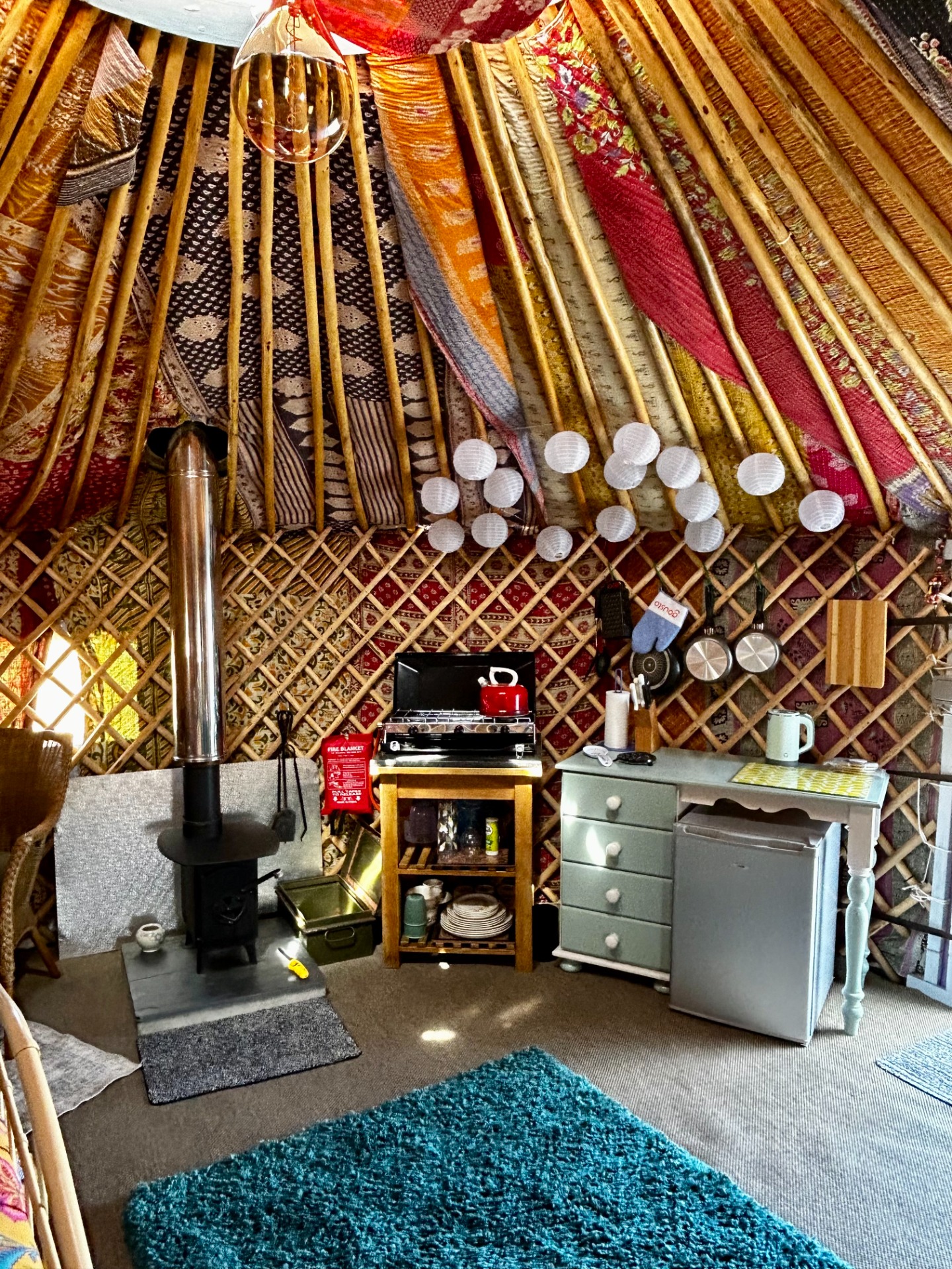 Cozy yurt interior with hanging lantern, striped rugs, wooden shelves, and a small stove and kettle.
